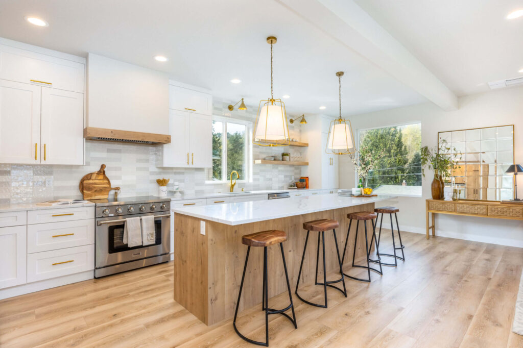 A bright modern kitchen with white shaker cabinets, gold hardware, and a large wood-accented island topped with white marble, paired with wooden barstools, pendant lights, stainless steel appliances, and light wood flooring, creating an airy and elegant design.