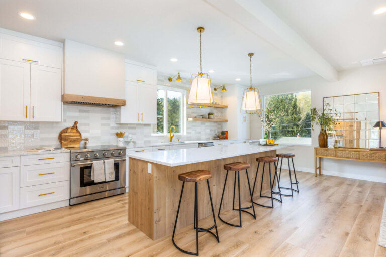A bright modern kitchen with white shaker cabinets, gold hardware, and a large wood-accented island topped with white marble, paired with wooden barstools, pendant lights, stainless steel appliances, and light wood flooring, creating an airy and elegant design.